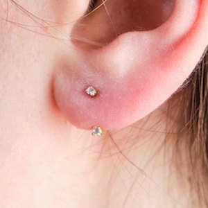 May include: Close-up of a person's ear with two small diamond stud earrings. The earrings are set in gold.