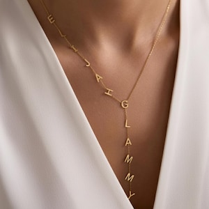 May include: A gold-colored necklace with the name "ELIJAH GLAMMY" spelled out in individual letters. The necklace has a delicate chain and a Y-shaped design, with the letters hanging down the front. The background is a white blouse.