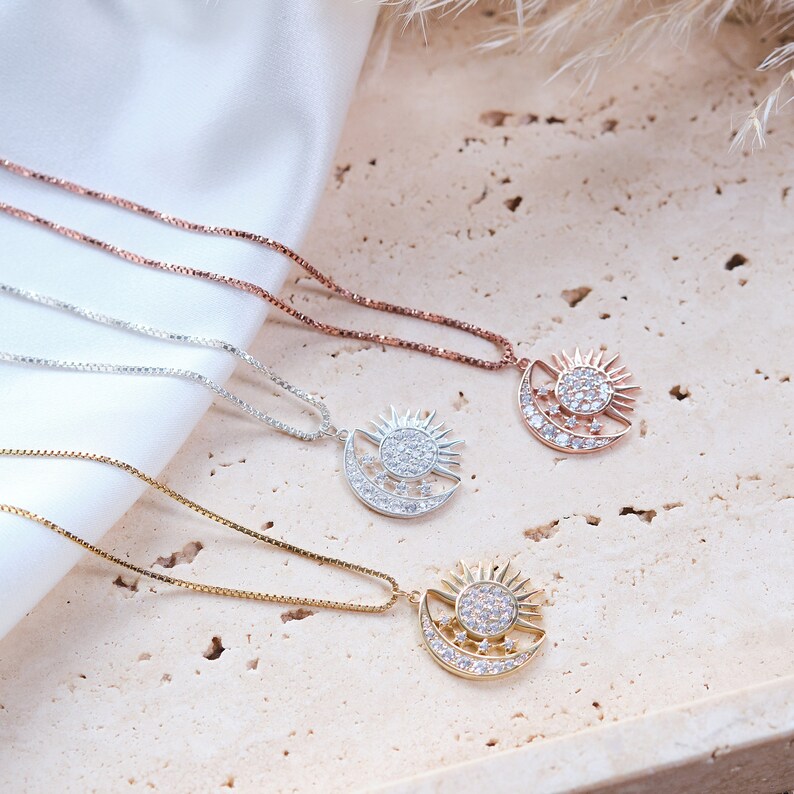 May include: Three silver necklaces with a sun and moon pendant. The pendants are adorned with sparkling clear stones. The necklaces are on a light brown textured surface.