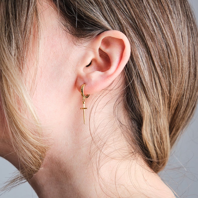 Peut inclure: Une boucle d'oreille en or avec un pendentif en forme de croix. La boucle d'oreille est port&eacute;e &agrave; l'oreille gauche d'une personne aux cheveux longs blonds.