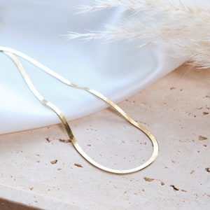 May include: A gold chain necklace with a flat, snake-like design. The chain is lying on a light brown textured surface.