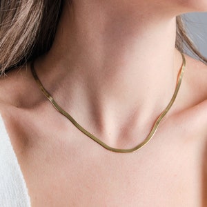 May include: A gold chain necklace with a flat, snake-like design. The necklace is worn by a person with a white shirt.