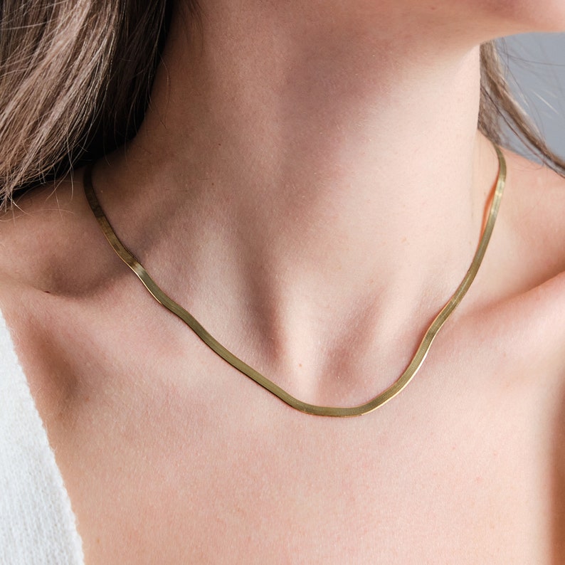 May include: A gold chain necklace with a flat, snake-like design. The necklace is worn by a person with a white shirt.