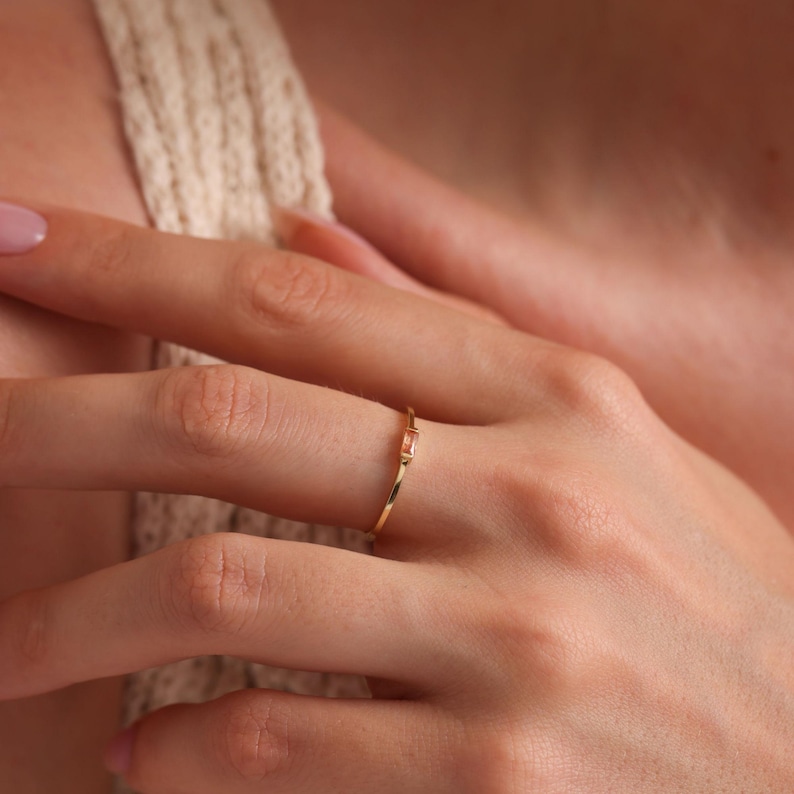 May include: A delicate gold ring with a single, small, rectangular, light pink gemstone.