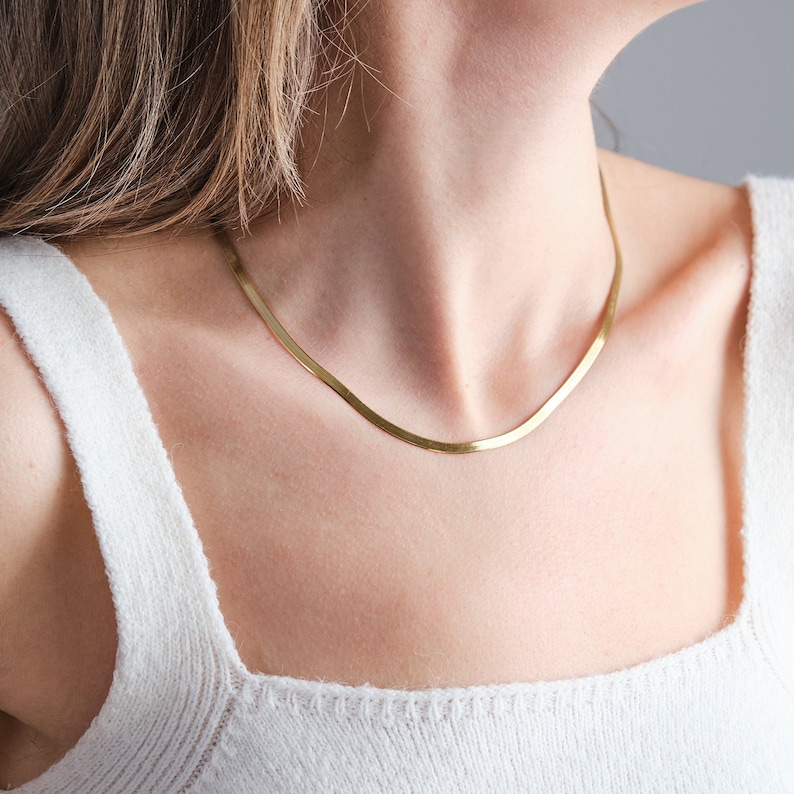 May include: A gold chain necklace with a simple, flat design. The chain is worn by a person with long brown hair.