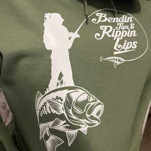 Pode incluir: Uma camisola de capuz verde com uma silhueta branca de uma pessoa a pescar em cima de um baixo. O texto "Bendin Tips & Rippin Lips" est&aacute; num c&iacute;rculo branco com um isco de pesca branco.