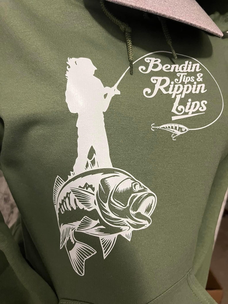 Pode incluir: Uma camisola de capuz verde com uma silhueta branca de uma pessoa a pescar em cima de um baixo. O texto "Bendin Tips & Rippin Lips" est&aacute; num c&iacute;rculo branco com um isco de pesca branco.