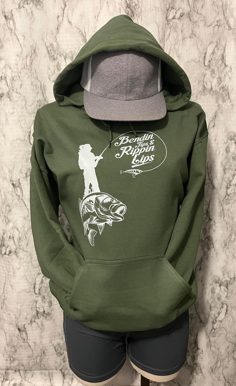 Pode incluir: Uma camisola de capuz verde com um gr&aacute;fico branco de uma pessoa a pescar em cima de um baixo. O gr&aacute;fico tamb&eacute;m inclui o texto "Bendin Tips & Rippin Lips".