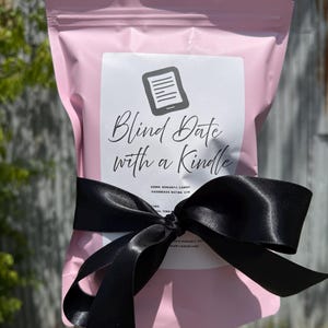 May include: A pink pouch with a white label featuring a Kindle icon and the text "Blind Date with a Kindle." The pouch is tied with a black satin ribbon. The label also includes the genre and Goodreads rating.