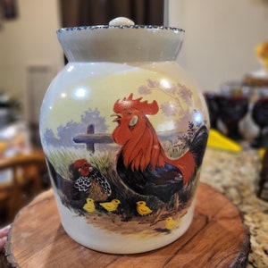 May include: A ceramic rooster cookie jar with a white lid and a rooster and chicks design. The rooster is red, black, and white, and the chicks are yellow. The jar is sitting on a wooden base.