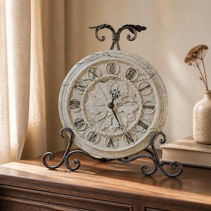 May include: A round table clock with a cream-colored face featuring dark green marble-like veining, black Roman numerals, and ornate central relief detailing. The clock is supported by a black metal stand with scrollwork and a top finial.