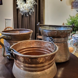 May include: Three copper pots with handles, two with iron handles and one with a copper handle. The pots are on a dark wood surface.