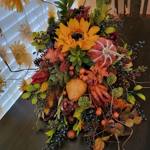 May include: A decorative floral arrangement featuring a large yellow sunflower, a red and white striped pumpkin, a yellow pear, green succulents, pinecones, dark blue berries, and various autumn-colored leaves and stems arranged in a dense cluster.