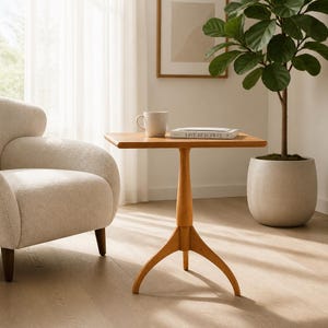 May include: A light brown wooden side table with a square top and a central pedestal base featuring three curved legs. A white ceramic mug and a book with the text "LIVE BEAUTIFUL" on the cover are placed on the flat surface of the table.