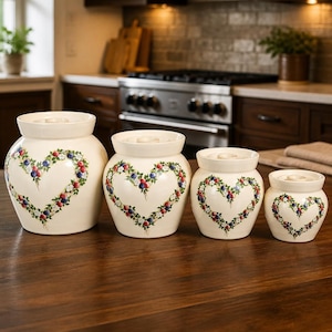 May include: A set of four cream-colored ceramic canisters with matching lids, arranged in descending order of size. Each canister features a hand-painted heart-shaped wreath design composed of green leaves, small white flowers, and red and blue berries.