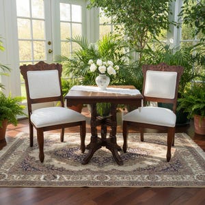 May include: A square wooden table with a central pedestal base and two matching chairs featuring dark brown wood frames and off-white upholstered seats and backrests. A white lace runner and a white floral vase sit on the table surface.