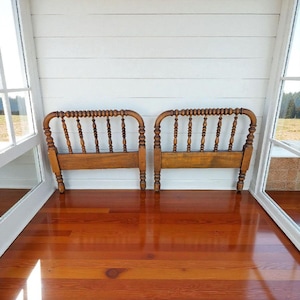May include: Two antique wooden headboards with a spindle design. The headboards are a warm brown color and stand on short legs. They are positioned against a white paneled wall, with a glossy wooden floor reflecting the light.