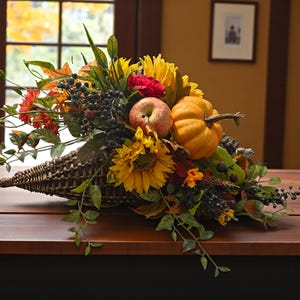 May include: A woven cornucopia overflowing with a harvest arrangement. The display includes sunflowers, a red rose, an apple, a small pumpkin, and berries. Green leaves and foliage add to the autumnal theme. The arrangement sits on a wooden surface.