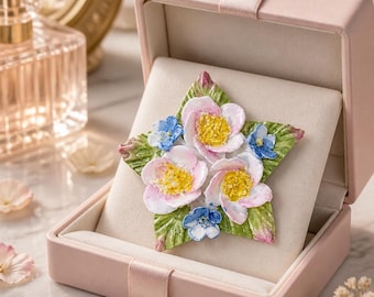 May include: A star-shaped brooch featuring sculpted pink and blue flowers with textured yellow centers and green leaves. The brooch is displayed inside an open, light pink rectangular jewelry box with a soft, cream-colored velvet interior lining.
