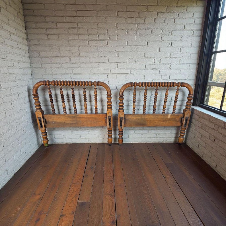 May include: Two antique wooden headboards with a warm, natural finish. Each headboard features a curved top with vertical spindles and decorative turned posts. The headboards are set against a white brick wall and a wooden floor.