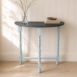 May include: A semi-circular console table with a dark gray top and three light blue turned legs. A clear glass vase with white floral branches sits on the left side, and a small brass dish rests on two stacked books on the right side.