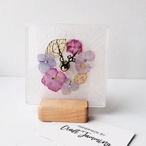 May include: A white square clock with a floral design made of pressed flowers and gold leaf. The clock is on a wooden stand and has a black face with gold hands. The clock is handmade by Craft Jamming.