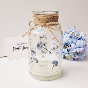 May include: A clear glass jar with a brown twine bow and a blue bead. The jar is filled with dried blue flowers and glitter. The jar is sitting on a white surface. The text "Handmade by Craft Jam" is on a white card.