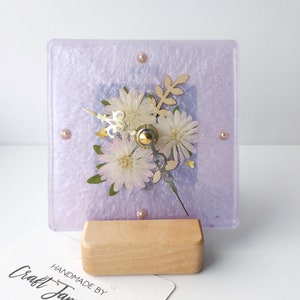 May include: A square, lavender glass clock with a floral design and a wooden stand. The clock face features pressed flowers and leaves. The clock is handmade by Craft Jamming.