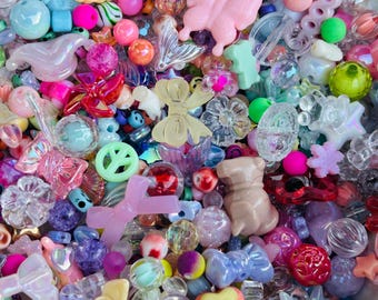 Puede incluir: Un surtido colorido de cuentas y abalorios artesanales de diversas formas y tamaños. La mezcla incluye mariposas, lazos, flores y corazones en tonos rosa, azul, amarillo y transparente. Las cuentas son de plástico y vidrio.