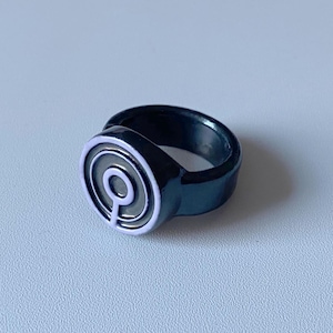 May include: A black ring with a circular design. The ring features a silver-colored inset with a stylized, concentric circle pattern. The ring is set against a plain, light-colored background.