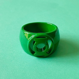 May include: A glossy, bright green ring featuring the Green Lantern symbol. The ring is a solid, rounded shape with a circular emblem on the front. The ring is set against a solid, light green background.