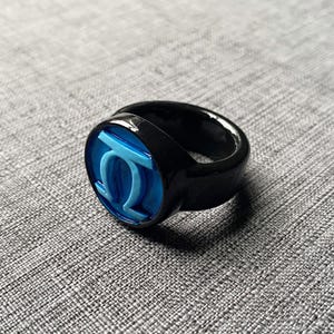 May include: A black ring with a blue and white emblem. The ring has a glossy finish and a thick band. The emblem features a stylized design in a circular setting. The ring is set against a gray textured background.