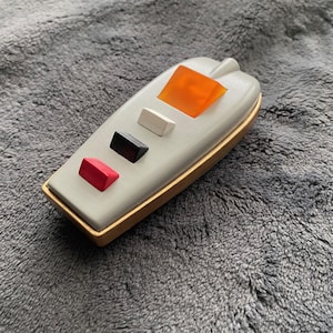 May include: A gray, boat-shaped object with a gold trim and colorful accents. It features a red, black, and white rectangular button, and an orange, trapezoid-shaped piece. The object is likely a decorative item or a unique piece of art.