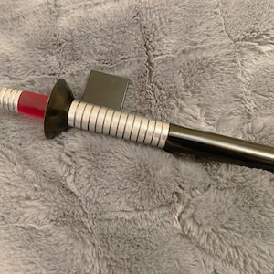 May include: A black and silver toy lightsaber with a red blade. The hilt has a black handle and a silver grip with vertical lines. A black rectangular piece is attached to the hilt.