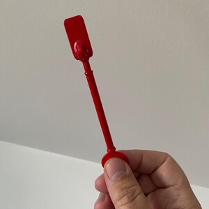 May include: A bright red cocktail muddler with a rectangular head and a long, slender handle. The muddler is held against a white background. The muddler is a bar tool used to mash ingredients in cocktails.