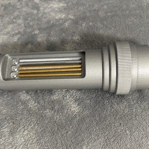 May include: A silver-colored cylindrical tool with a rectangular opening revealing several parallel rods in silver and gold tones. The tool has a ridged section and a black angled tip. The object is lying on a gray, textured surface.