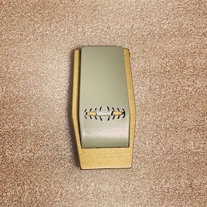May include: A rectangular money clip with a light green center and gold-colored edges. The center section has a decorative stitched design. The clip is shown against a textured, light brown background.