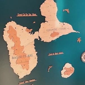 May include: A wooden map of Guadeloupe, a French island in the Caribbean Sea. The map is mounted on a teal blue background. The map is divided into two main islands, Grande-Terre and Basse-Terre. The map also includes the smaller islands of Marie-Galante, Saintes, and Saint-Martin. The map is labeled with the names of the islands and other geographical features. The map is decorated with small wooden cutouts of sea turtles and dolphins.