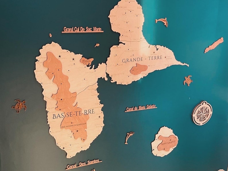 May include: A wooden map of Guadeloupe, a French island in the Caribbean Sea. The map is mounted on a teal blue background. The map is divided into two main islands, Grande-Terre and Basse-Terre. The map also includes the smaller islands of Marie-Galante, Saintes, and Saint-Martin. The map is labeled with the names of the islands and other geographical features. The map is decorated with small wooden cutouts of sea turtles and dolphins.