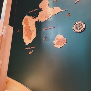 May include: A green wall with a wooden map of the islands of Guadeloupe. The map includes the islands of Grande Terre, Basse Terre, Marie Galante, and the surrounding islands. The map is surrounded by wooden cutouts of fish and a compass.