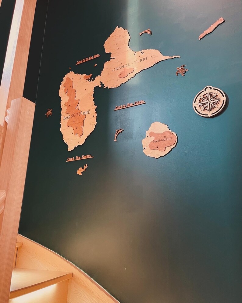 May include: A green wall with a wooden map of the islands of Guadeloupe. The map includes the islands of Grande Terre, Basse Terre, Marie Galante, and the surrounding islands. The map is surrounded by wooden cutouts of fish and a compass.