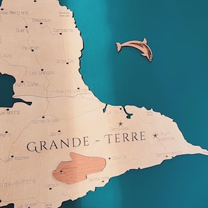 May include: A wooden map of the island of Guadeloupe, with the words 'GRANDE - TERRE' printed in black. The map is mounted on a teal blue wall. The map shows the names of various cities and towns on the island, including Pointe-a-Pitre, Le Gosier, and Saint-Francois.