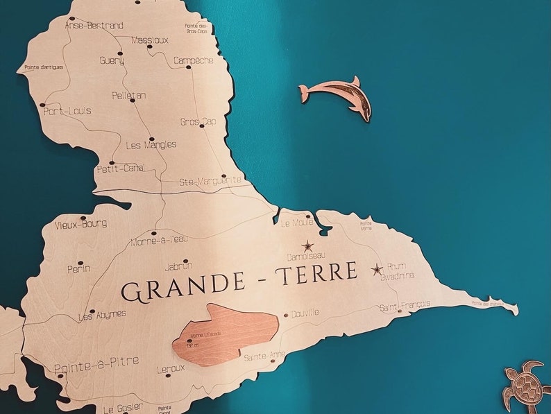May include: A wooden map of the island of Guadeloupe, with the words 'GRANDE - TERRE' printed in black. The map is mounted on a teal blue wall. The map shows the names of various cities and towns on the island, including Pointe-a-Pitre, Le Gosier, and Saint-Francois.