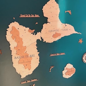 May include: A teal blue background with a wooden map of Guadeloupe. The map is divided into two islands, Basse-Terre and Grande-Terre. The map is labeled with the names of the islands and the surrounding bodies of water. The map is surrounded by small wooden cutouts of sea turtles, dolphins, and a compass.