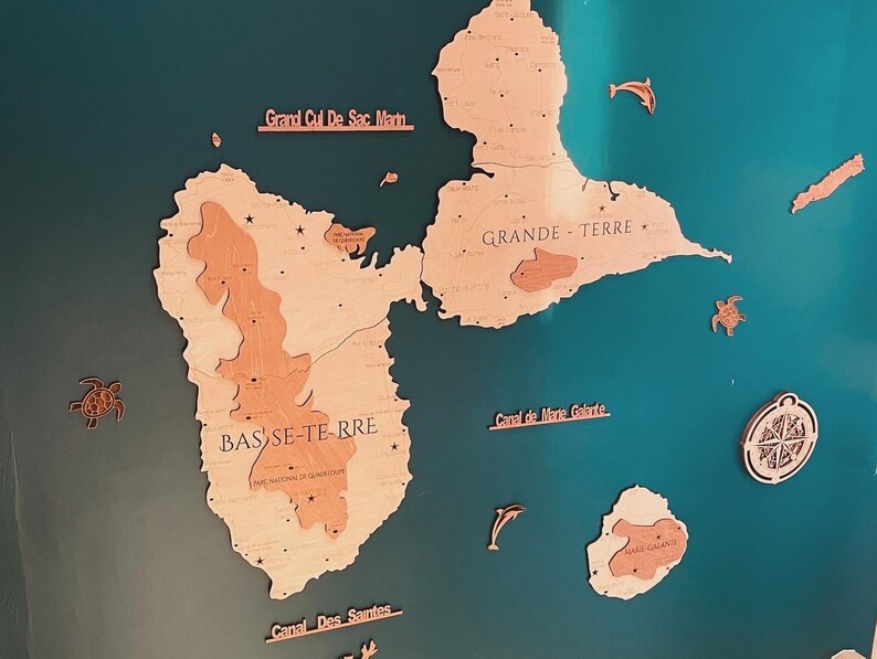May include: A teal blue background with a wooden map of Guadeloupe. The map is divided into two islands, Basse-Terre and Grande-Terre. The map is labeled with the names of the islands and the surrounding bodies of water. The map is surrounded by small wooden cutouts of sea turtles, dolphins, and a compass.