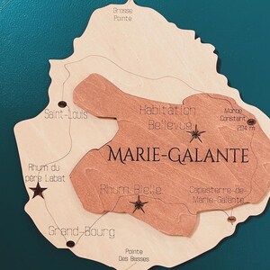 May include: A wooden map of Marie-Galante, a French island in the Caribbean. The map is brown and beige and features the names of various locations on the island, including Grosse Pointe, Saint-Louis, Habitation Bellevue, Morne Constant, Rhum du pere Labat, Rhum Bielle, Grand-Bourg, Pointe Des Basses, and Capesterre-de-Marie-Galante. The map is surrounded by a teal background.