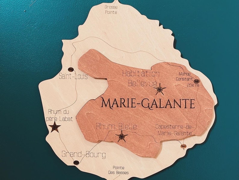 May include: A wooden map of Marie-Galante, a French island in the Caribbean. The map is brown and beige and features the names of various locations on the island, including Grosse Pointe, Saint-Louis, Habitation Bellevue, Morne Constant, Rhum du pere Labat, Rhum Bielle, Grand-Bourg, Pointe Des Basses, and Capesterre-de-Marie-Galante. The map is surrounded by a teal background.