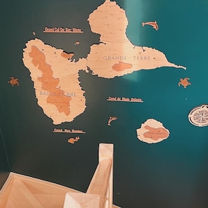 May include: A wooden map of the islands of Guadeloupe, with the names 'Grand Cul De Sac Marm', 'GRANDE TERRE', 'BAS SE-TE RRE', 'Canal de Mane Galante', and 'Canal Des Santes' written on it. The map is mounted on a teal wall. The map is surrounded by wooden cutouts of sea turtles, dolphins, and a compass.