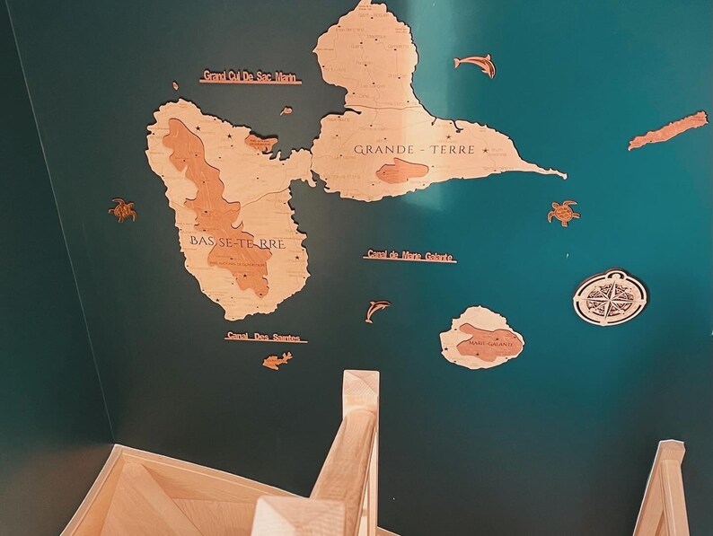 May include: A wooden map of the islands of Guadeloupe, with the names 'Grand Cul De Sac Marm', 'GRANDE TERRE', 'BAS SE-TE RRE', 'Canal de Mane Galante', and 'Canal Des Santes' written on it. The map is mounted on a teal wall. The map is surrounded by wooden cutouts of sea turtles, dolphins, and a compass.