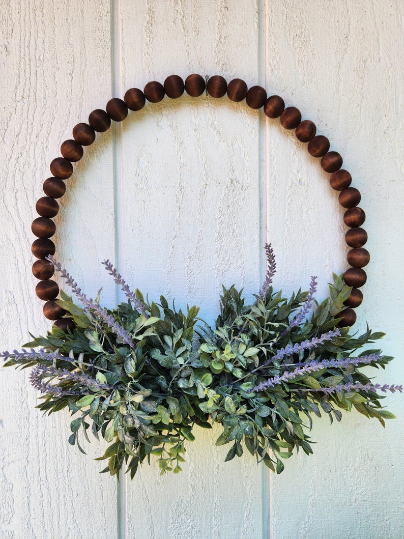 May include: A brown wooden bead wreath with a green and purple floral arrangement. The wreath is hanging on a white wooden wall.